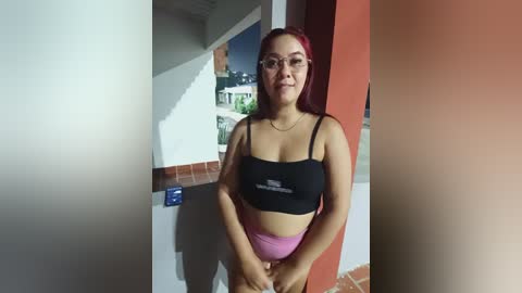 Video of a young woman with long red hair, wearing glasses and a black sports bra with pink shorts, standing indoors near a red wall.