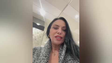 Video of a light-skinned woman with long black hair, wearing a grey and white knit cardigan and a necklace, standing in a modern bathroom with beige walls and a mirror.