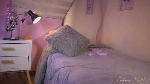 Video of a cozy, pastel-colored bedroom with a lavender bed, textured throw pillows, a white nightstand, a black desk lamp, and a wooden floor.
