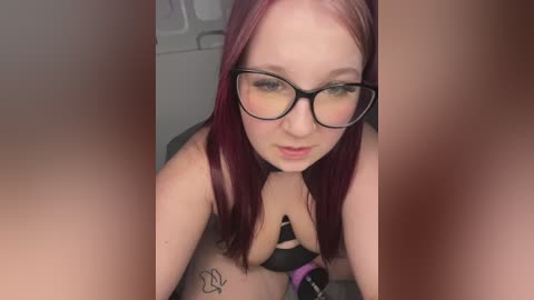 A video of a fair-skinned, red-haired woman with glasses and black lingerie, crouching on a tiled floor. She has a butterfly tattoo on her left hip. The background is blurred.