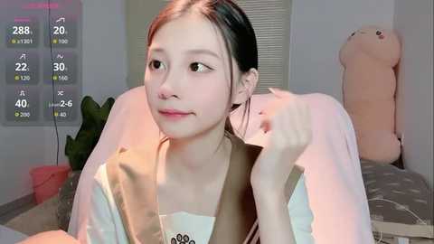 A video of an Asian woman with fair skin, straight black hair, and a small nose, wearing a beige jacket, sitting indoors with a teddy bear.