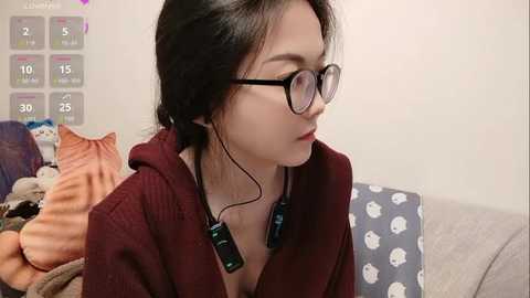 Video of a young Asian woman with glasses, dark hair, wearing a burgundy robe, seated on a couch with stuffed animals. Background includes a calendar and a blue pillow with white polka dots.