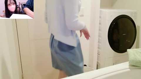 A video of a woman in a white sweater and blue shorts standing in a beige-tiled bathroom with a washing machine and a white sink. A close-up of another woman with dark hair is in the upper left corner.