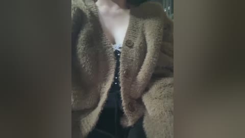 Video of a person wearing a beige, chunky-knit cardigan over a black, sequin dress with a plunging neckline, partially visible. The background is blurred, creating a soft focus effect.