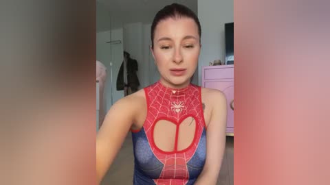 A video of a fair-skinned woman with brown hair in a ponytail, wearing a tight, red and blue Spider-Man costume with a cutout heart on the chest. She is indoors, in a bedroom with light-colored walls, a pink dresser, and a dark figure in the background.