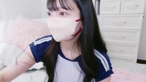 A video of an Asian woman with long black hair, wearing a blue and white sports shirt and a white face mask, standing in a bright, modern bedroom with white furniture and pink bedding.