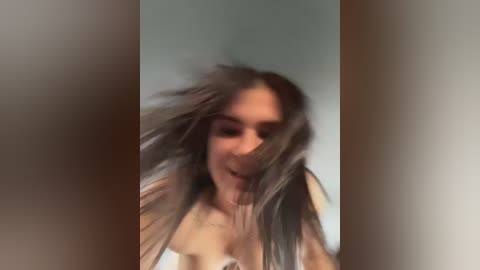 Video of a young woman with long, tousled dark hair, wearing a white top, smiling, with a blurred, out-of-focus background.