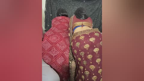 A video captures two women, one in a red saree with intricate white designs, the other in a maroon saree with golden floral patterns, lying on a bed with a black and white patterned bedspread. The setting is indoors with a warm, muted background.