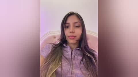 A video of a young Latina woman with long, straight, dark brown hair, wearing a purple checkered shirt, sitting against a light pink, tufted headboard.