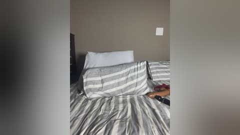 Video of a neatly made bed with gray and white striped bedding, a white pillow, and a small red object on the side, in a minimalist beige room with a white light switch on the wall.
