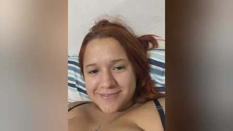 Media: Video of a young woman with fair skin, red hair, and a slight smile, lying on a bed with blue and white striped pillows, wearing a navy bra.