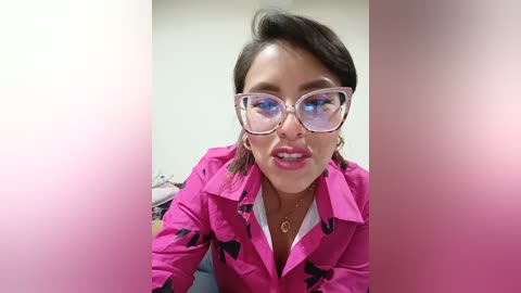 Video of a woman with fair skin, brown hair, and pink glasses, wearing a pink jacket with black designs, pink lipstick, and gold necklace. Background is blurred.