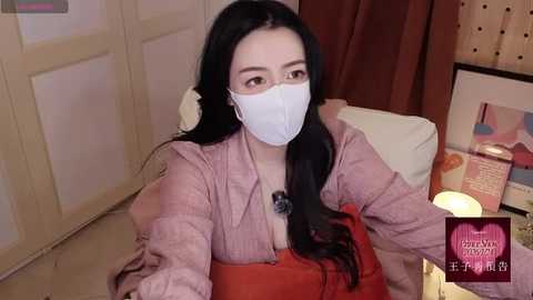 Video of an East Asian woman with long black hair, wearing a white face mask, pink cardigan, and orange bag, sitting indoors. Background includes a wooden door, soft lighting, and decor.