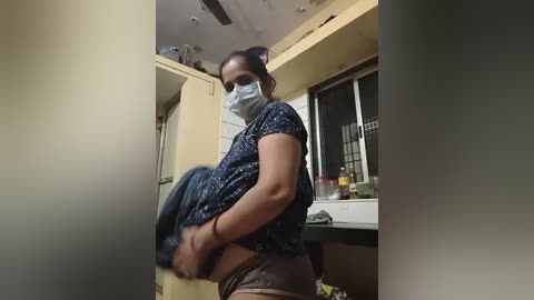 Video of a woman with dark skin, wearing a blue mask, blue patterned shirt, and black underwear, lifting a blue shirt in a small kitchen.
