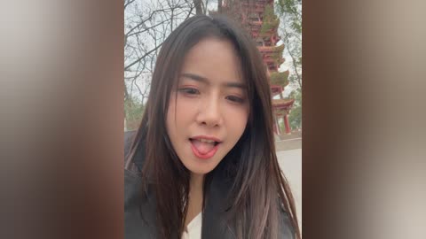 Video of an Asian woman with long black hair, wearing a black leather jacket, sticking out her tongue playfully outdoors, surrounded by traditional red and gold pagodas and leafless trees.