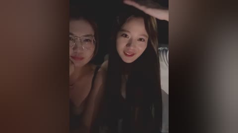 Video of two young Asian women, one with glasses, standing side by side, smiling warmly. They wear casual, dark clothing, framed by dim lighting and a dark background.
