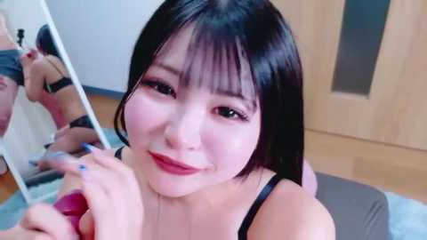 Video of a young Asian woman with straight black hair and pale skin, wearing a black bra, sitting on a gray cushion, looking at herself in a mirror, smiling.