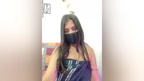 Video of a person with long, dark hair, wearing a black mask and a black strapless dress with white stripes. Background shows a white wall and a colorful patterned bedspread.