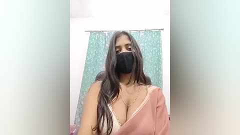 Video of a South Asian woman with long dark hair, wearing a pink satin blouse, black face mask, and a gold necklace, in a bathroom with a teal shower curtain.