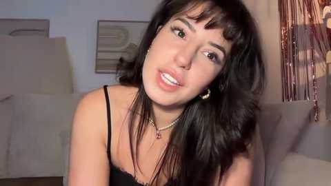 Video of a young woman with long black hair, fair skin, and dark eyes, wearing a black tank top, smiling, standing indoors with a white bed and metallic decor in the background.