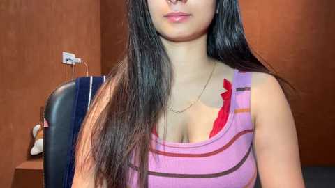 Video of a young woman with long black hair, fair skin, wearing a pink striped tank top that reveals a red bra underneath. She is indoors against a brown wall.