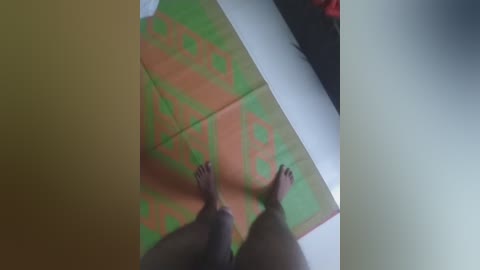 Video of a person's bare feet on a green and orange geometric-patterned mat, viewed from above. The background shows a white surface, possibly a bed or floor, with a hint of a dark object.