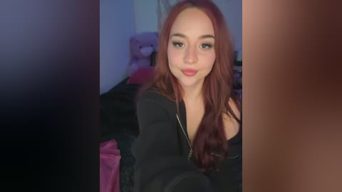 Video of a young woman with fair skin, long auburn hair, and full lips, wearing a black top. She has a confident expression. Background includes a pink bed and a teddy bear, in a dimly lit room.