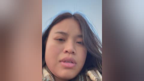Video of a young Asian girl with long, dark hair, wearing a patterned scarf. She has a neutral expression, with slightly puffy eyes, and her face is slightly blurred.