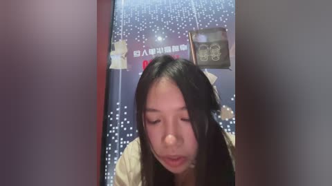 Video of an Asian woman with long black hair, wearing a beige sweater, looking down with a neutral expression, standing in front of a digital display screen with a dark background and white dots.