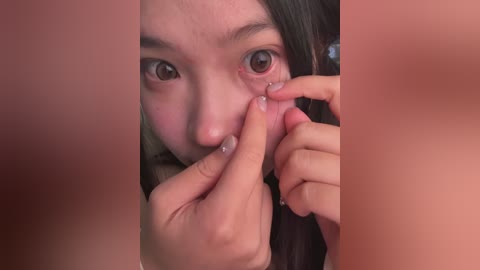 A close-up video of a young Asian woman with fair skin, dark eyes, and long black hair, gently pulling out a zit with her fingers. The image is blurred around the edges.