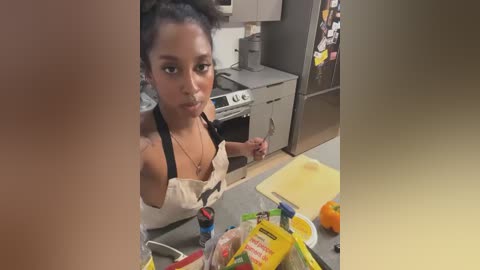 Video of a young Black girl with dark skin and curly hair, wearing a white apron, standing in a messy kitchen with a stove, fridge, and scattered food items.
