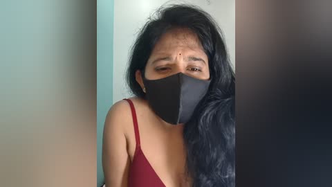 Video of an Asian woman with long black hair, wearing a black face mask and a red spaghetti-strap top, standing in front of a blurred background.