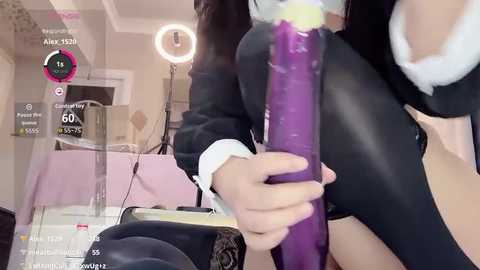 Video of a woman with light skin, holding a large, purple dildo, wearing a black fur-trimmed jacket and black tights, in a brightly-lit room with a pink background.