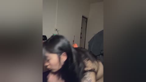 A blurred video shows a person with long black hair, possibly Asian, leaning against a wall, partially obscured by a blurred object. The background features a closed door and a room with minimal lighting.