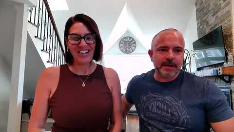 Video of a smiling woman with glasses and short brown hair in a sleeveless maroon top, and a bald man in a blue t-shirt, standing indoors with a wooden staircase and clock wall behind them.