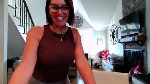 Video of a smiling woman with short brown hair and glasses, wearing a sleeveless brown top, standing in a modern living room with wooden floors, a fireplace, and decorative clocks on the wall.