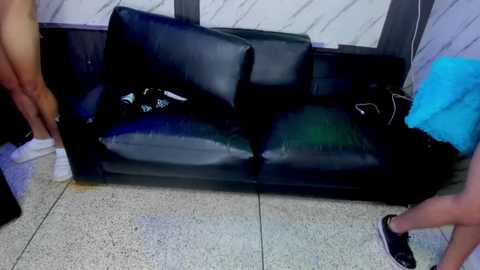 Video of a black leather couch in a tiled room with a white wall, showing a man in a blue shirt and another man in a black shirt standing near it.