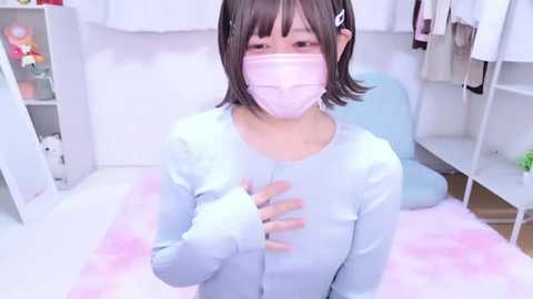 Video of an Asian woman with short dark hair, wearing a pink face mask, light blue cardigan, and white shirt, sitting on a pink rug in a white room with shelves and stuffed animals.