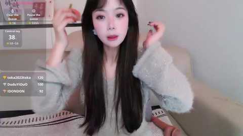 Video of an East Asian woman with long black hair, fair skin, and red lipstick, wearing a light gray sweater, sitting on a beige couch, with a digital overlay showing social media stats.