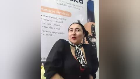 Video of a Latina woman with medium skin tone and dark hair in a bun, wearing a black blazer, patterned scarf, and red lipstick, standing in front of a poster promoting a medical conference.