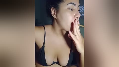 Video of a light-skinned woman with curly hair, wearing a black bra, sensually biting her finger, in a dimly lit room.