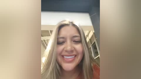 Video of a young Caucasian woman with long, straight blonde hair, fair skin, and a bright smile. She wears a red top. The background shows a modern indoor setting with beige and white walls and large windows.