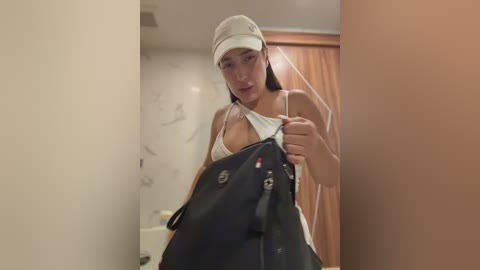 Video of a young Asian woman with straight black hair, wearing a white visor and a revealing white lace bra, holding a black trash bag, in a bathroom with beige walls and a wooden door.
