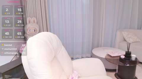 Video of a plush, white, tufted armchair in a modern living room with light gray curtains and a cream-colored couch. A digital display shows a \"Twitch\" live stream with user stats.