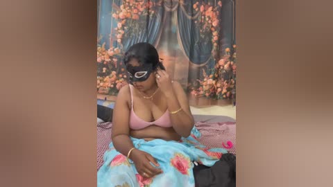 Video of a South Asian woman with medium-dark skin, wearing a pink bra and floral-patterned skirt, seated on a bed with a floral tapestry backdrop, holding a pink object in her hand.