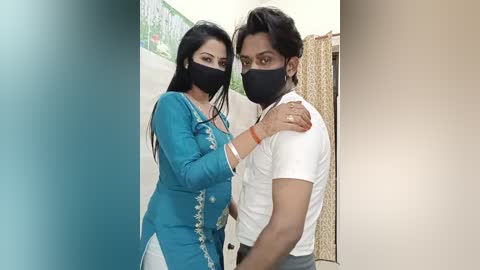 Video of two women wearing black face masks and traditional Indian outfits, standing closely together in a hallway with beige walls and green floral wallpaper.