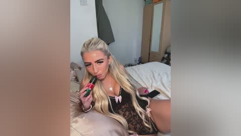 Video of a blonde woman with long, wavy hair, wearing black lingerie with pink bows, holding a green lipstick, lying on a bed in a bedroom.