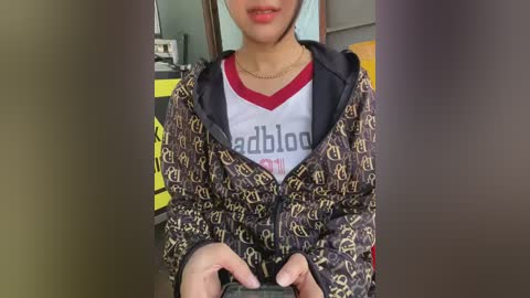 Video of a young Asian woman with light brown skin, wearing a dark, patterned jacket over a red and white t-shirt, holding a phone, indoors with blurred background.