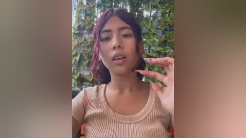 A video of a young woman with medium brown skin, wearing a beige ribbed tank top, posing against a lush green vine background. She has shoulder-length, wavy, maroon hair and is smiling with her hand near her mouth.