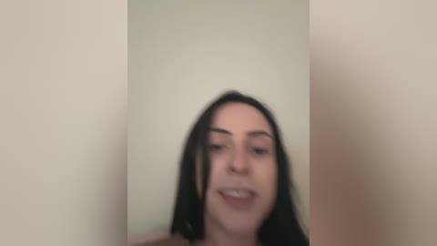 Media: Blurry video of a woman with straight black hair and fair skin, smiling, standing in a plain beige room with no visible clothing.